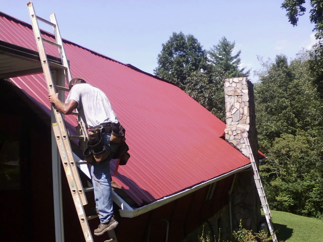Roofer on a ladder beside a red metal roof and stone chimney — Soffit & Fascia Repair by Roofing Brothers in Chillicothe